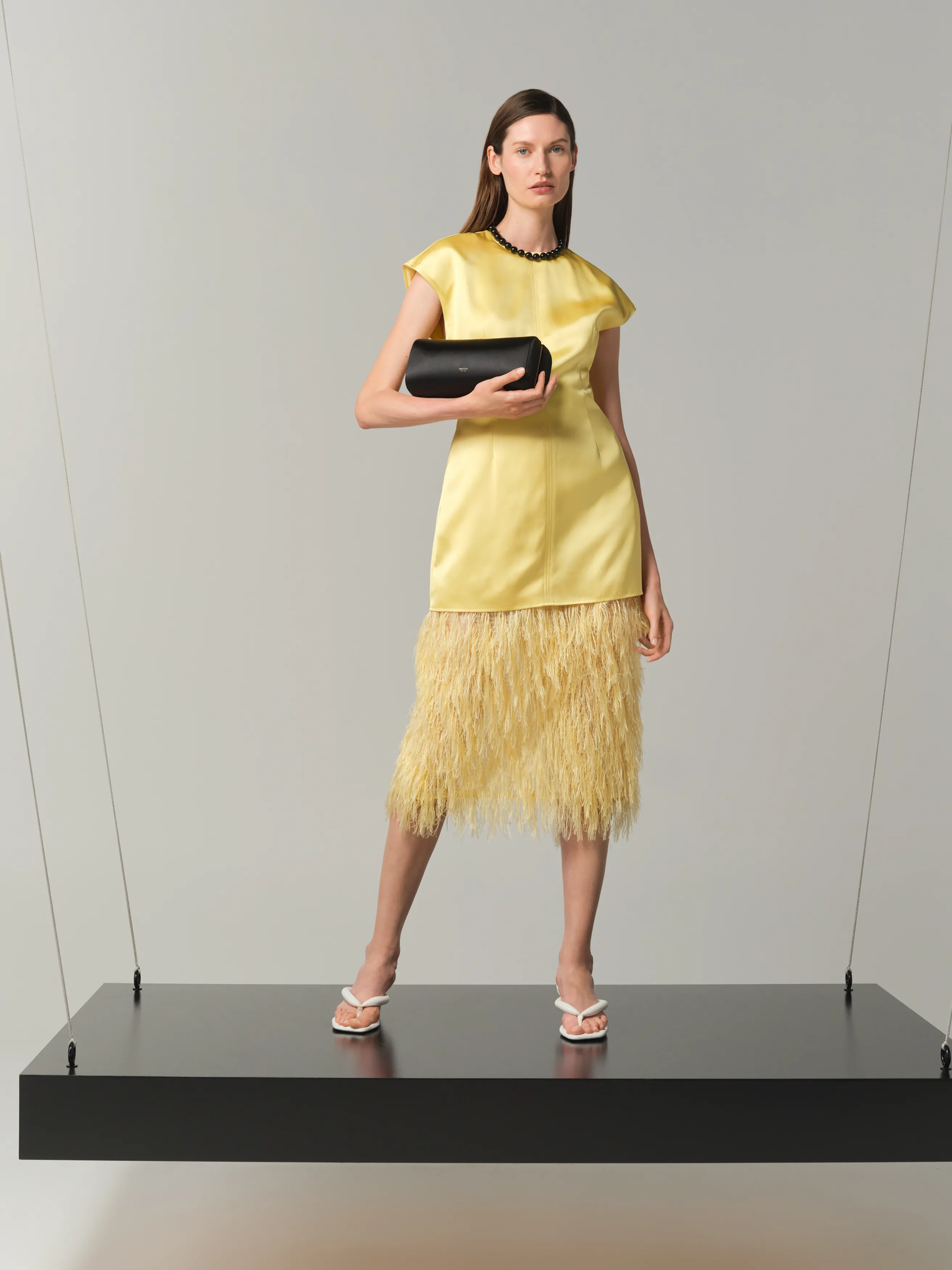 Model wearing a yellow dress with fringes, holding a black clutch.