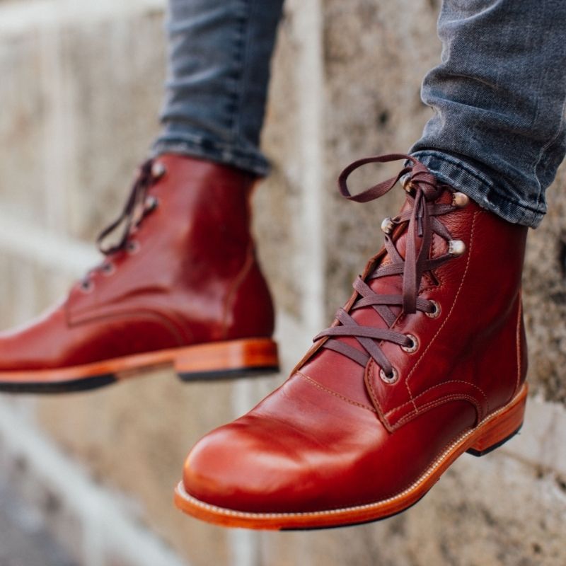 A man leaning against a stone wall wearing Adelante chestnut reddish-brown leather wide width lace-up boots.