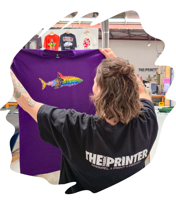 Staff member holding up purple t-shirt with colourful fish graphic, inspecting print clarity and alignment.