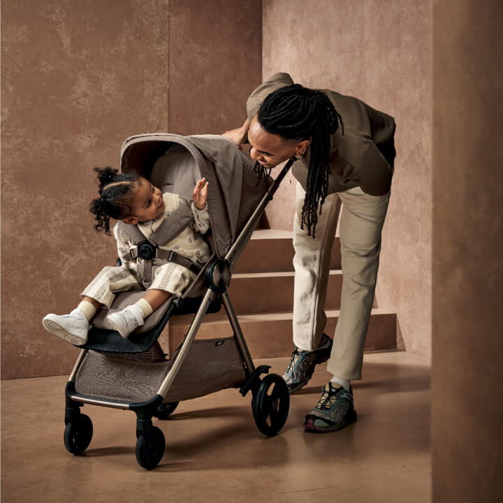 Adult interacting with a child in a stroller against a minimalist background.