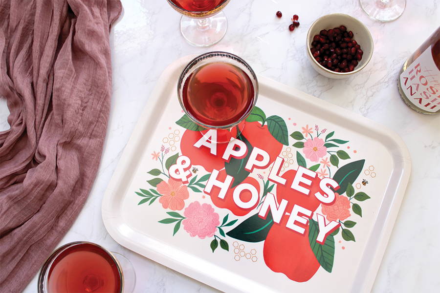 A serving tray that says apples and honey on it with champagne glasses of pomegranate bellinis.