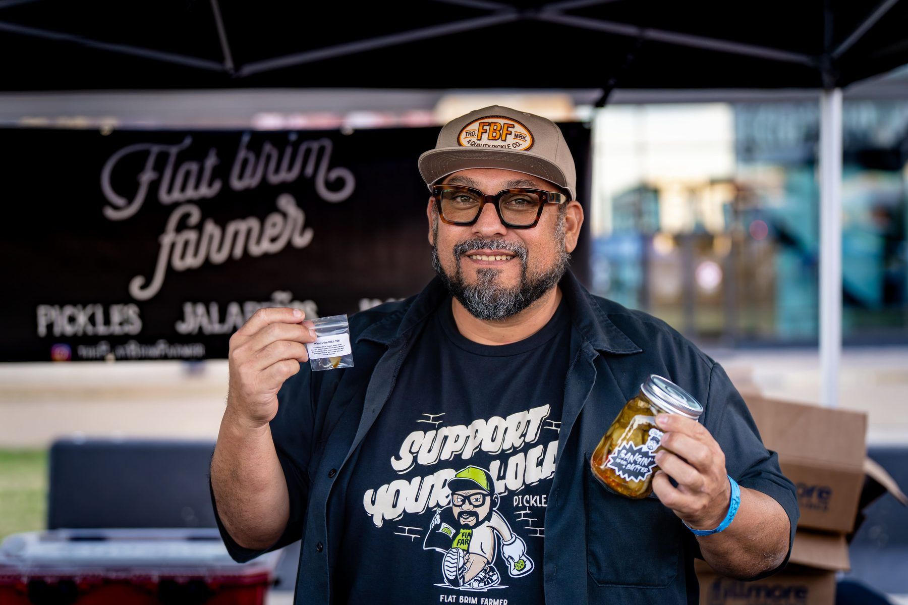 Flat Brim Farmer pickle vendor holding a sample and a jar of Bangin' Bread and Butter pickles at The Big Dill.