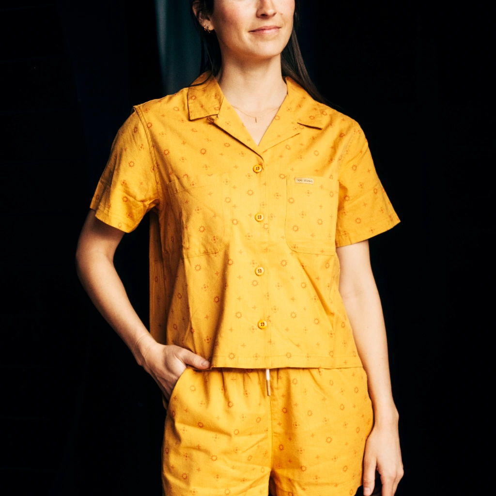 A woman in yellow pajamas stands against a dark background, smiling.