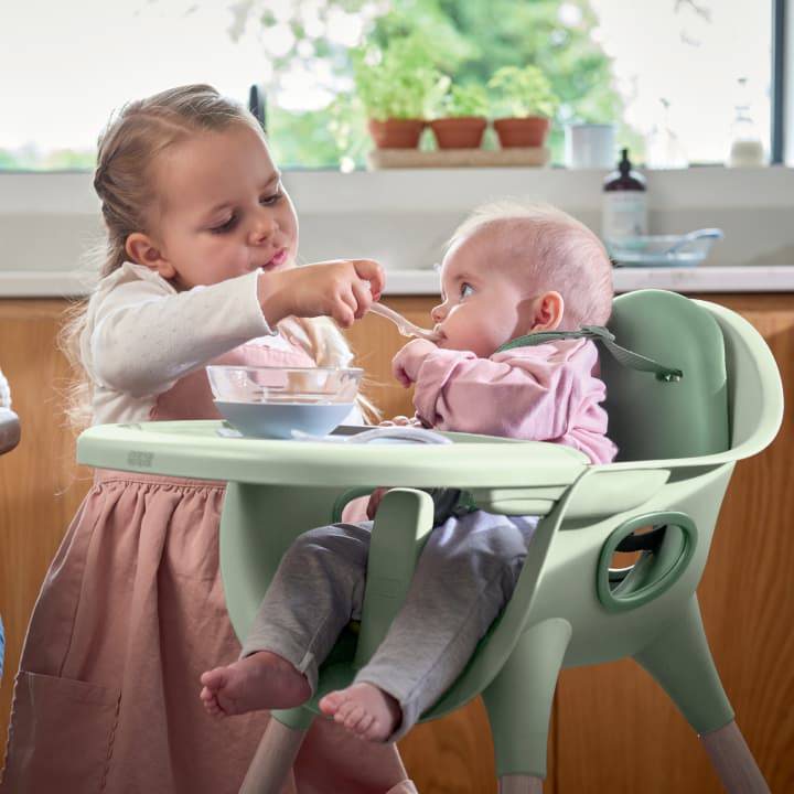 A baby girl is sitting in the Juice highchair being fed by her older sister.