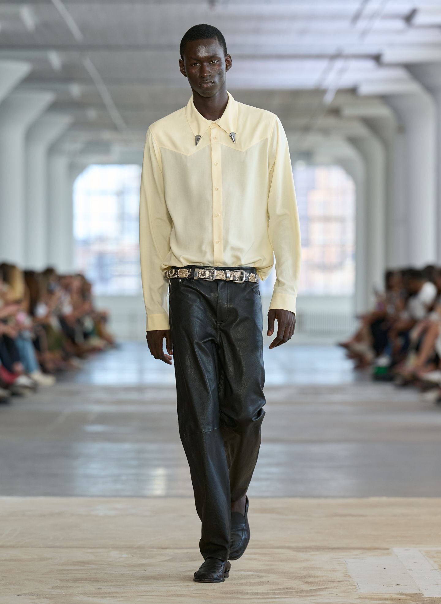 Model walking in a fashion show, wearing a yellow shirt and black pants.