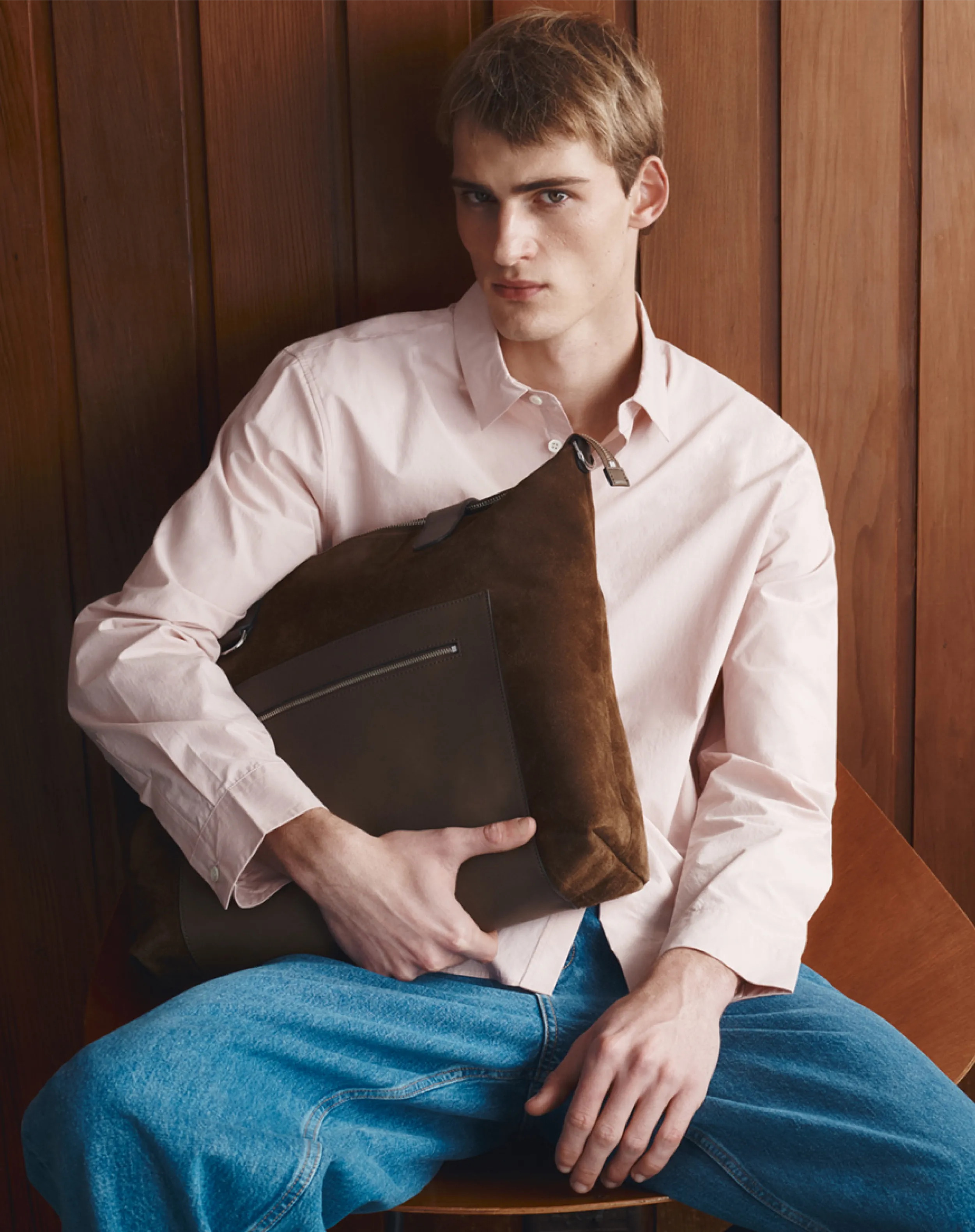 Young man in a pink shirt holding a brown bag, seated against wooden background.