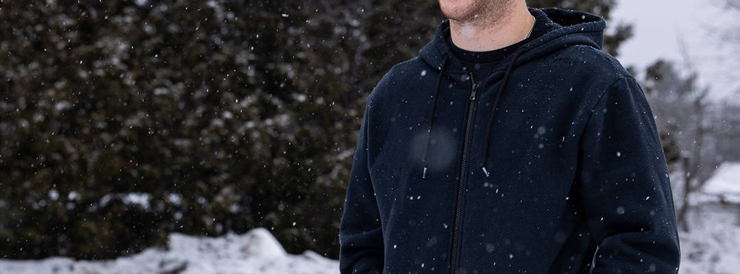 A man standing in falling snow wearing a blue hoodie.