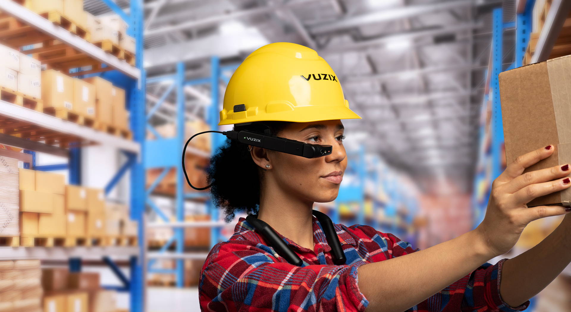 Young woman wearing Vuzix smart glasses mounted on a yellow hard hat scanning a box on a shelf in a warehouse.
