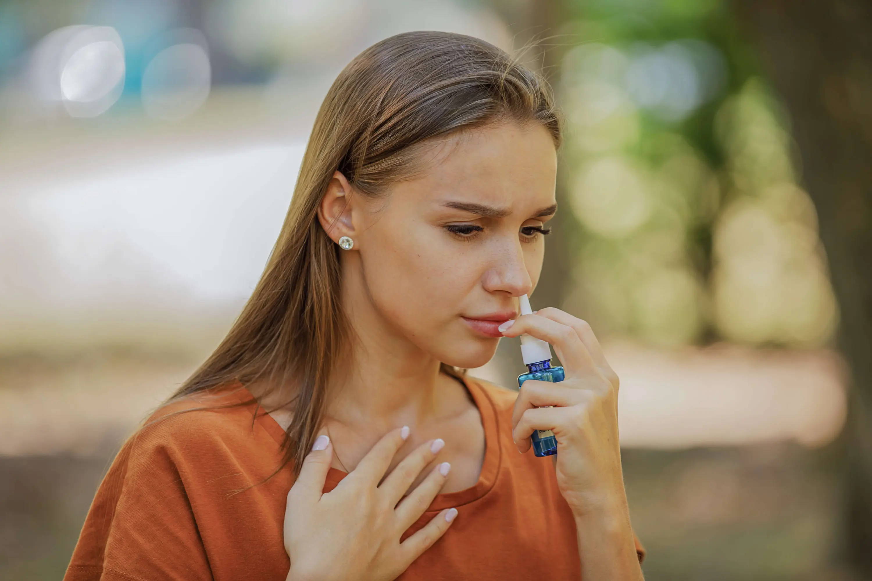Een vrouw met halflang bruin haar staat in een park en gebruikt neusspray omdat ze last heeft van allergieklachten. 