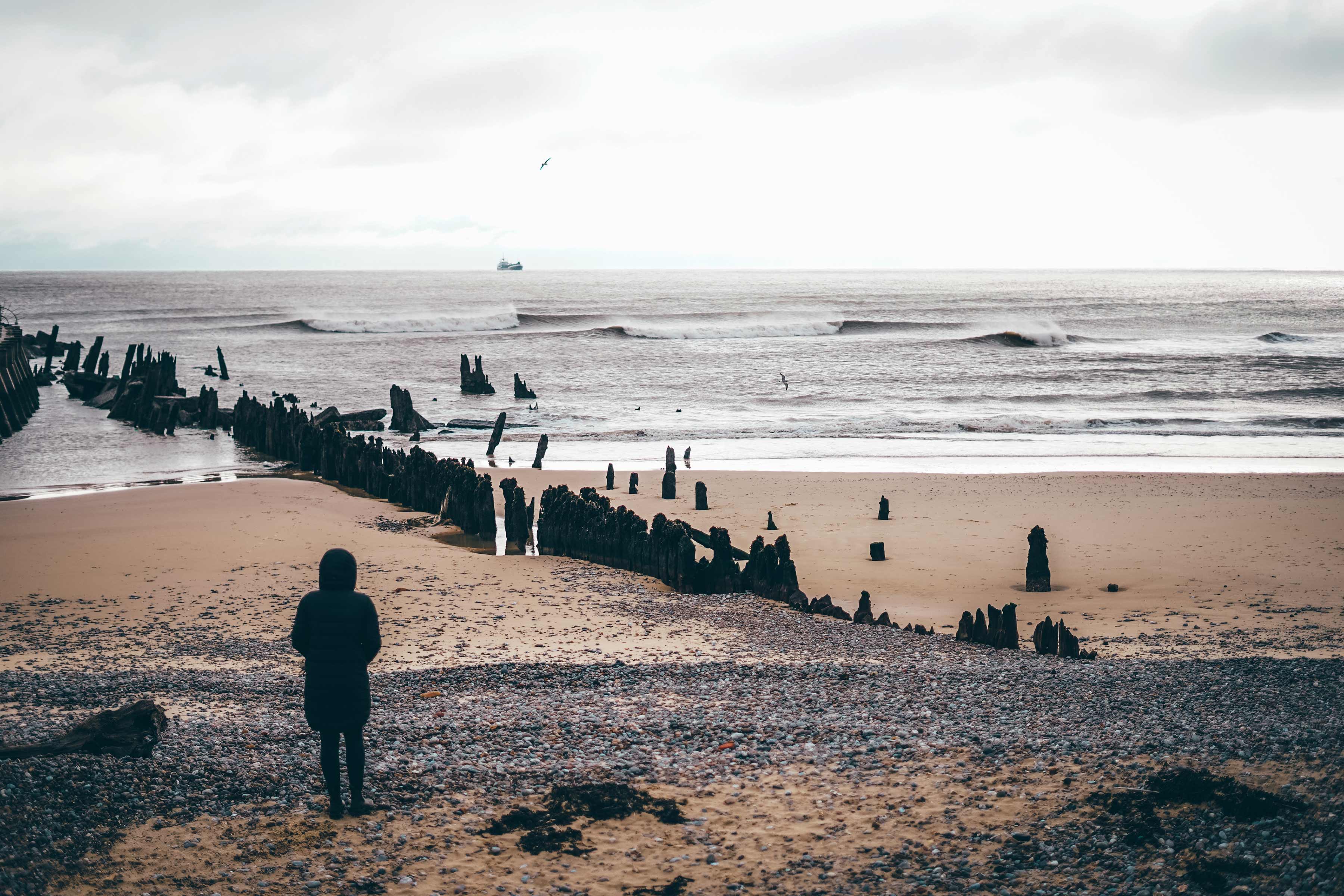 Person standing on beach, looking at waves and distant figures. Cloudy sky.