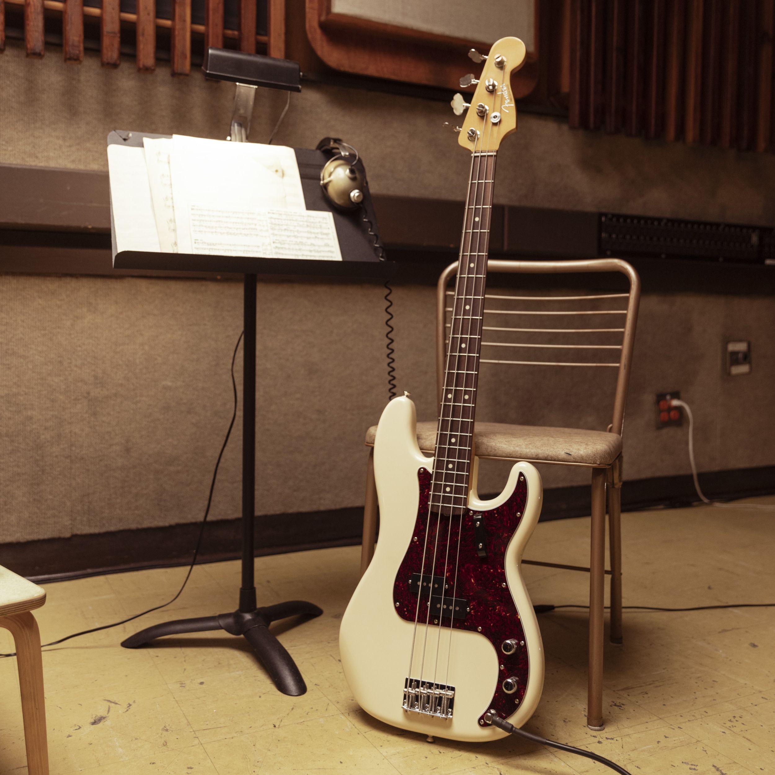 A white electric bass guitar beside a music stand and chair.