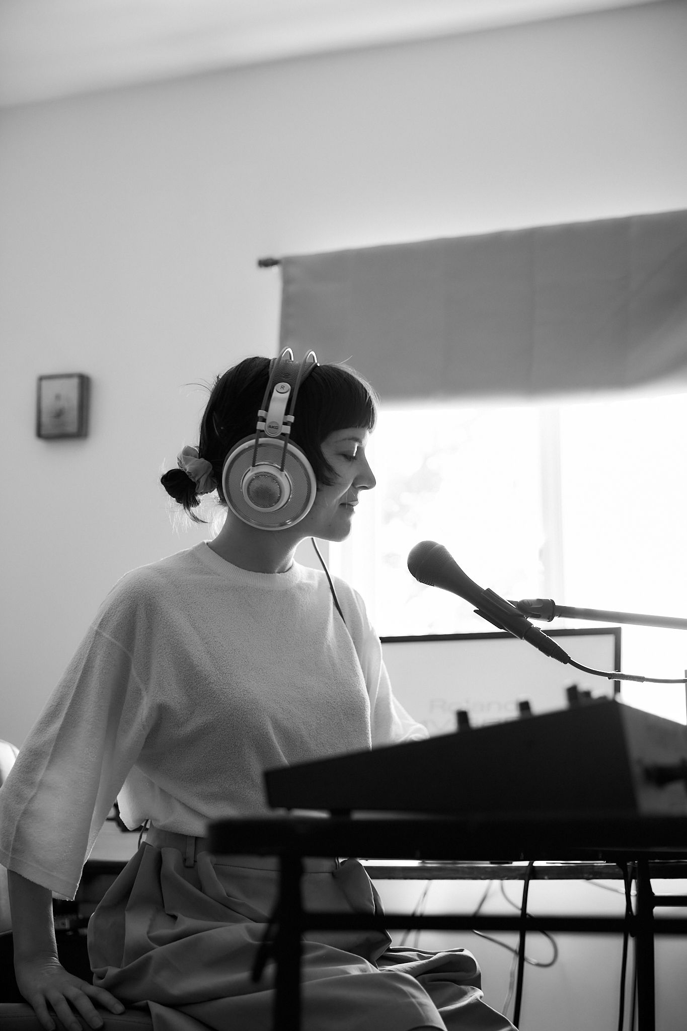 A woman wearing headphones sits at a keyboard in a bright room.