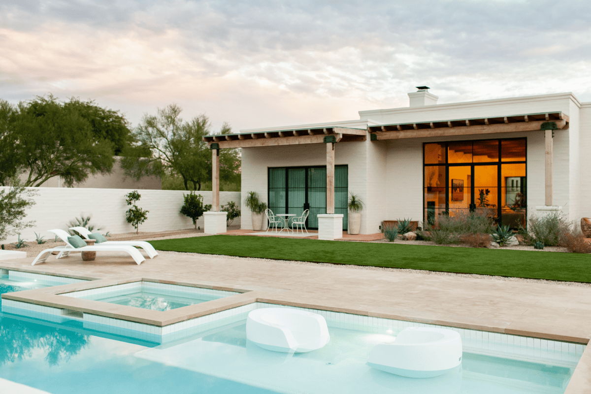 Modern pool patio with white in-pool chaise lounges, white pool floats, a small bistro set, and covered outdoor living space beside a rectangular swimming pool.