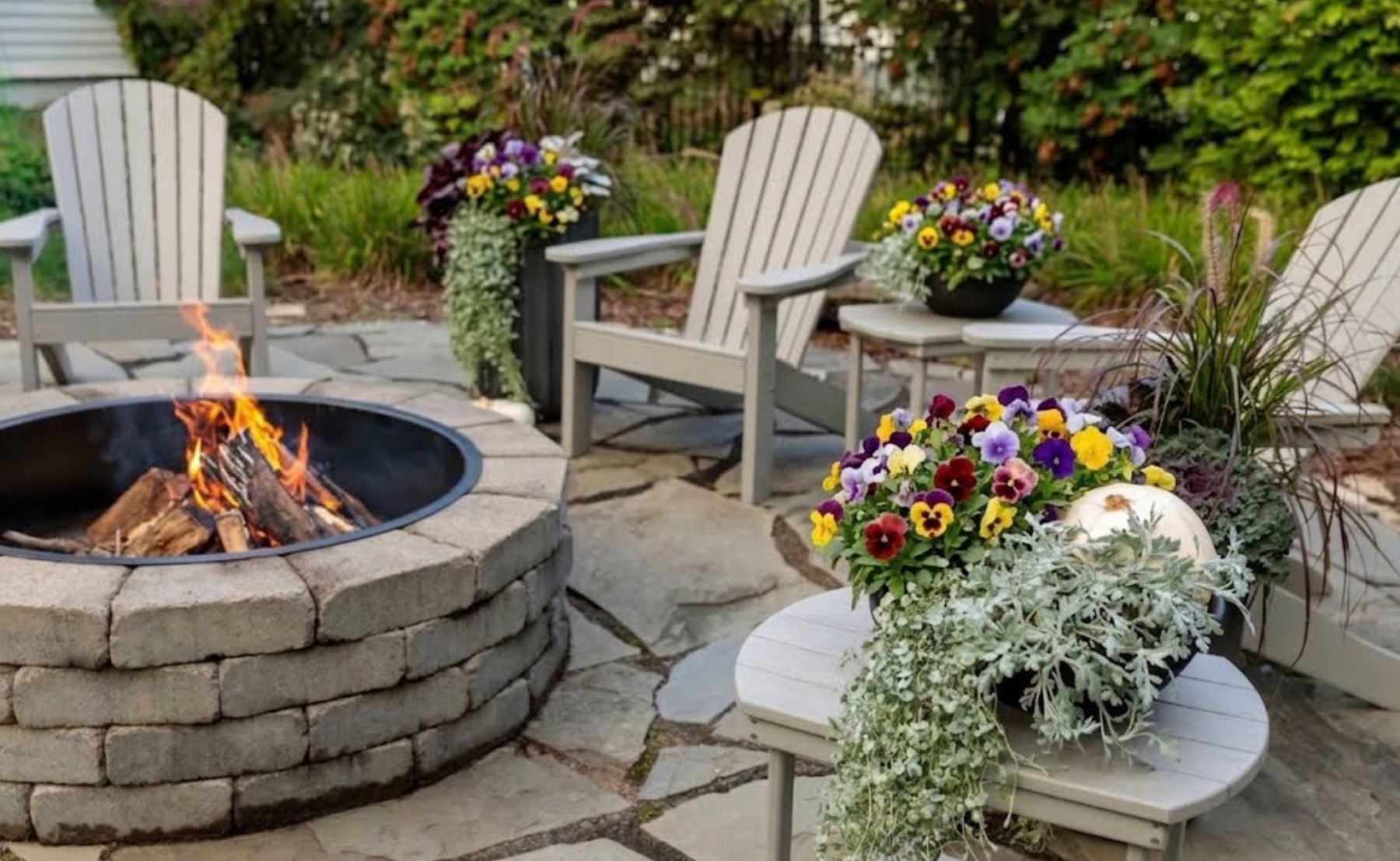 Scene of fall patio seating area featuring Adirondack chairs around a stone fire pit, surrounded by colorful pansies, ornamental grasses, and seasonal container plantings.