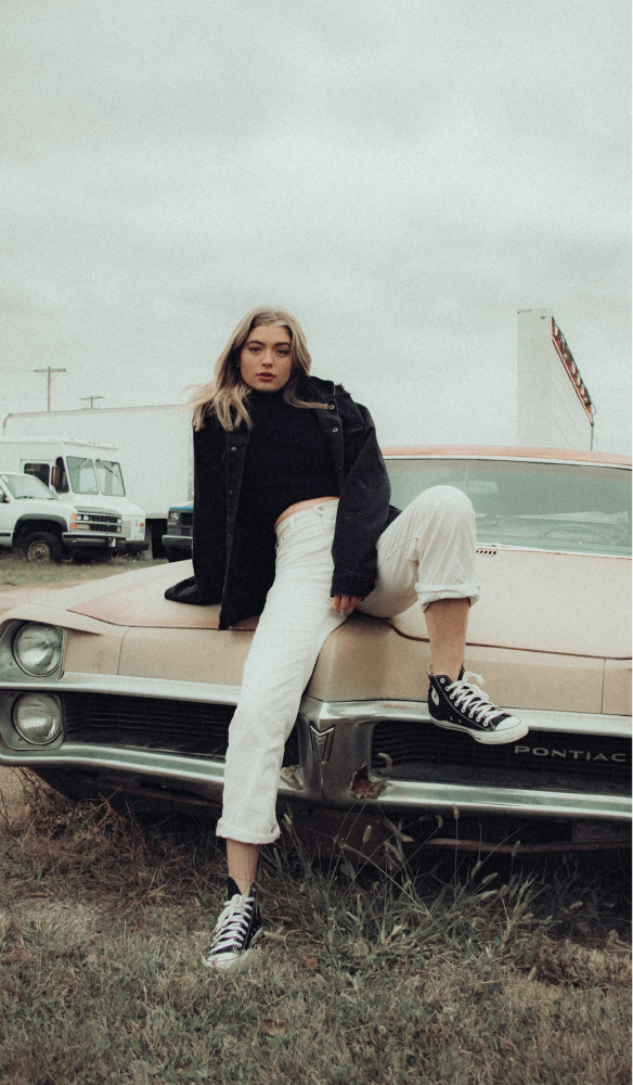A woman wearing white pants, a black jacket and converse sneakers sat on the hood of a classic car