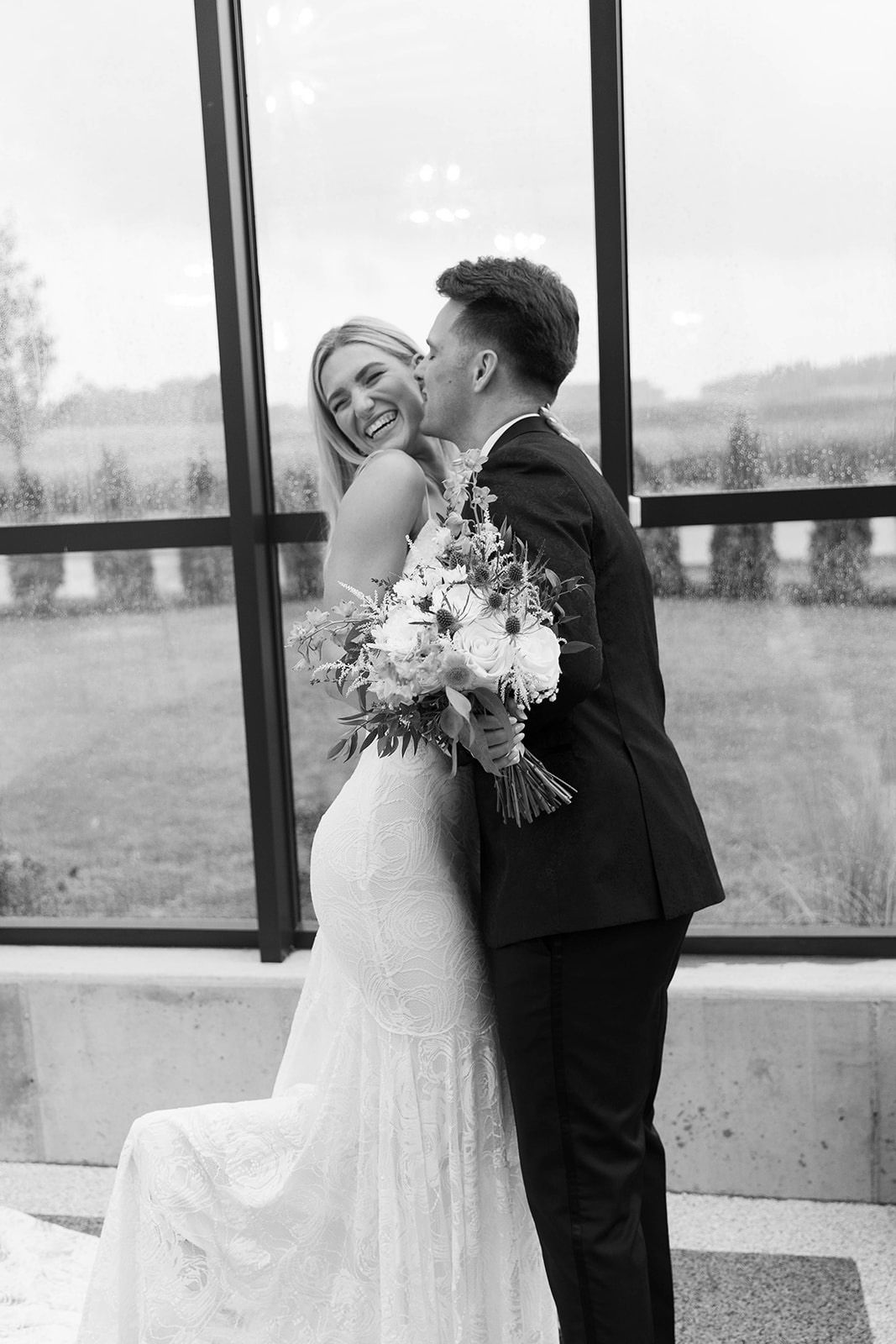 Couple embracing, bride holding bouquet, smiling in front of large window.