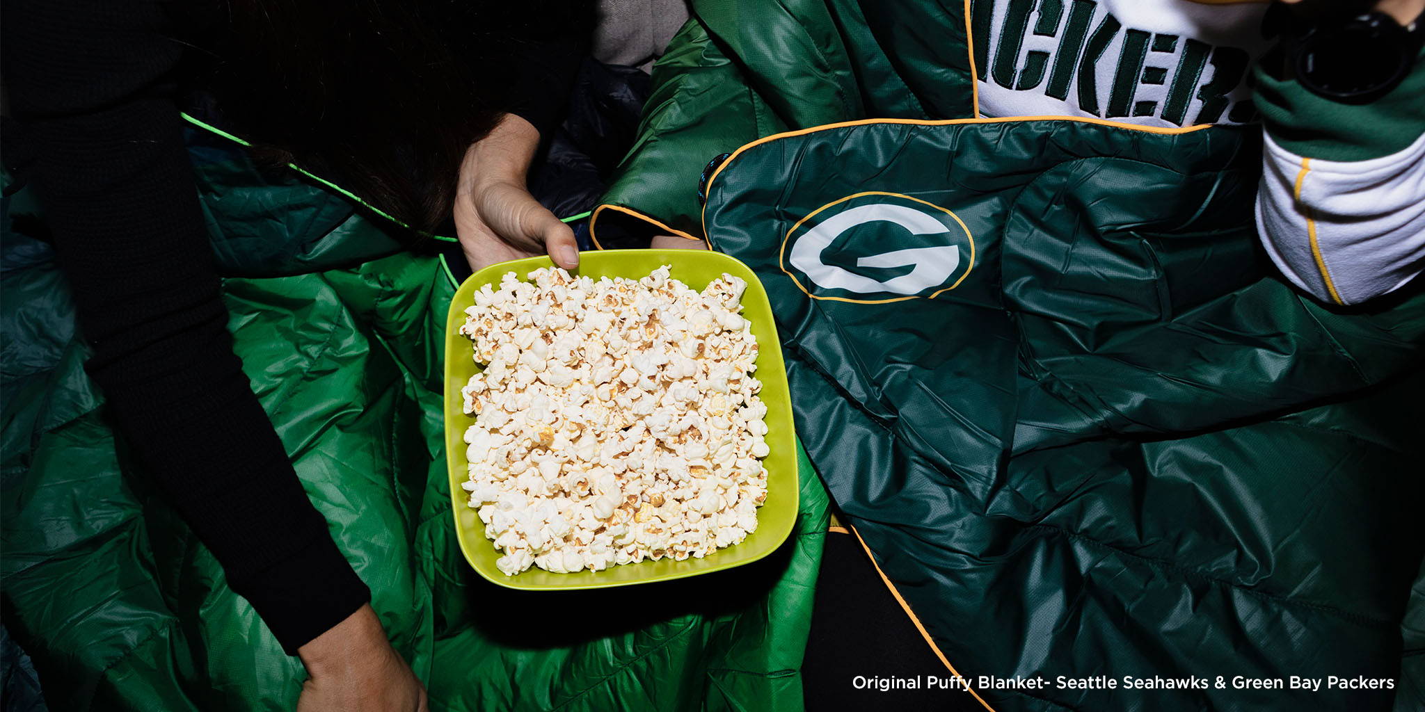 Two people wearing Rumpl NFL blankets with a bowl of popcorn