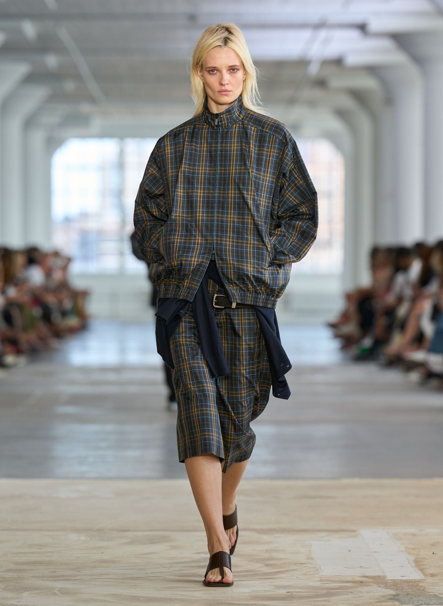 Model walking down runway wearing plaid outfit and sandals.