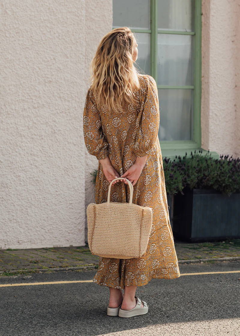 A model wearing a mustard coloured paisley printed dress holding a crochet shoulder bag behind her back 