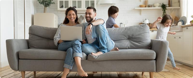 Smiling couple sitting on a couch using a laptop while two children play in the background