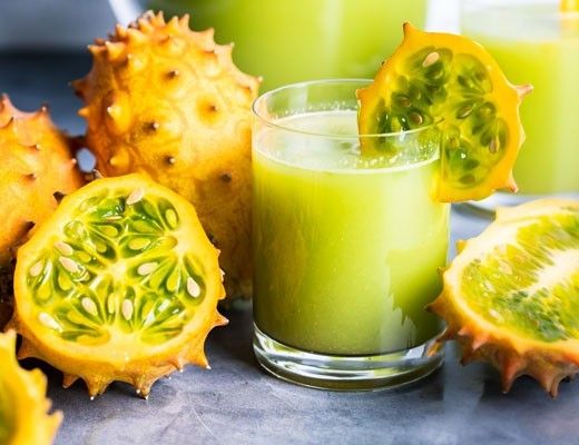 Glass of green juice surrounded by whole and sliced kiwano fruits.