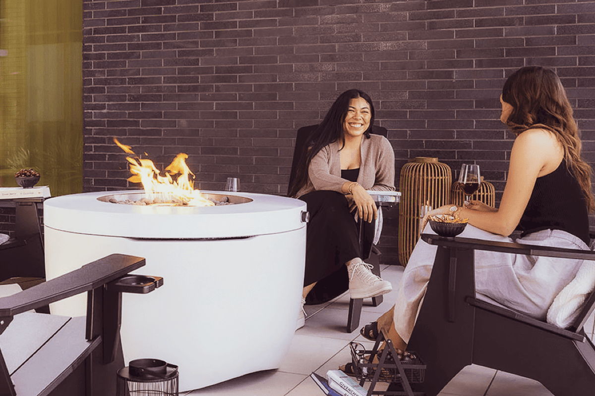 Two people talking beside a round outdoor fire pit in a modern patio conversation area.