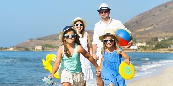 Family enjoying a beach day, wearing hats and sunglasses, holding toys.