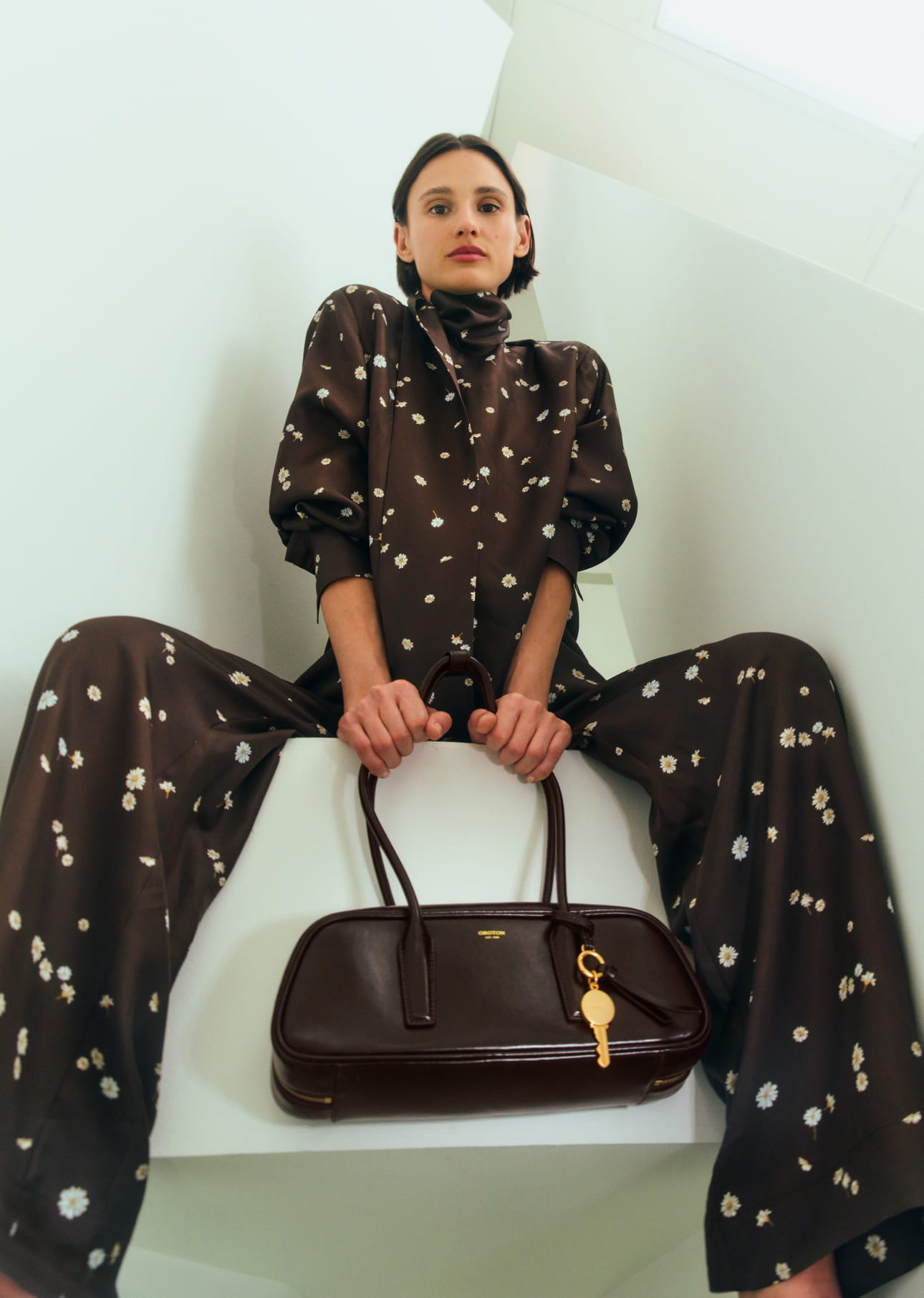 Model in patterned outfit holding a handbag, seated in a modern setting.