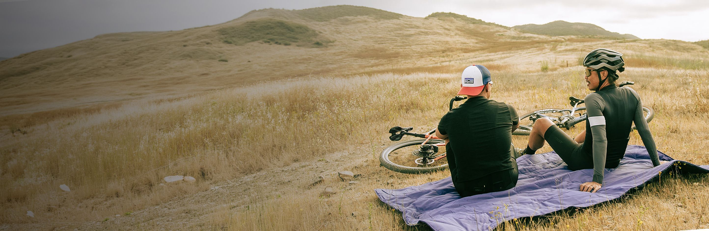 Two cyclists resting on a blanket in a hilly landscape.