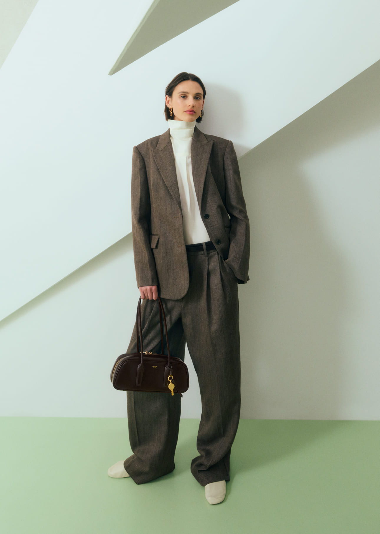 A model in an oversized brown suit holding a handbag against a minimalist background.