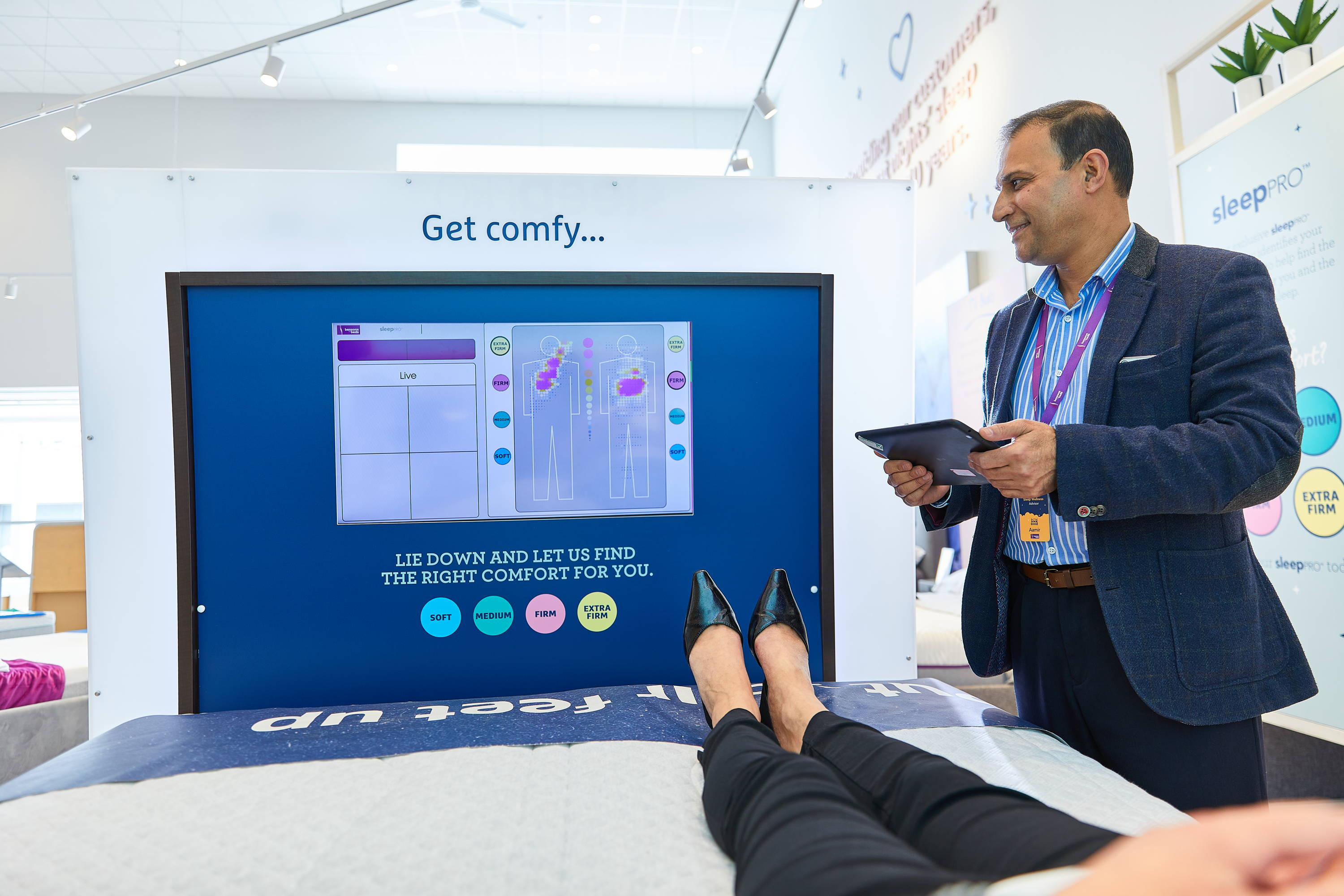 View of woman's trousered legs and shoes as she lies on a SleepPRO with the screen in view. Man stands beside the bed smiling and holding a computerised tablet.