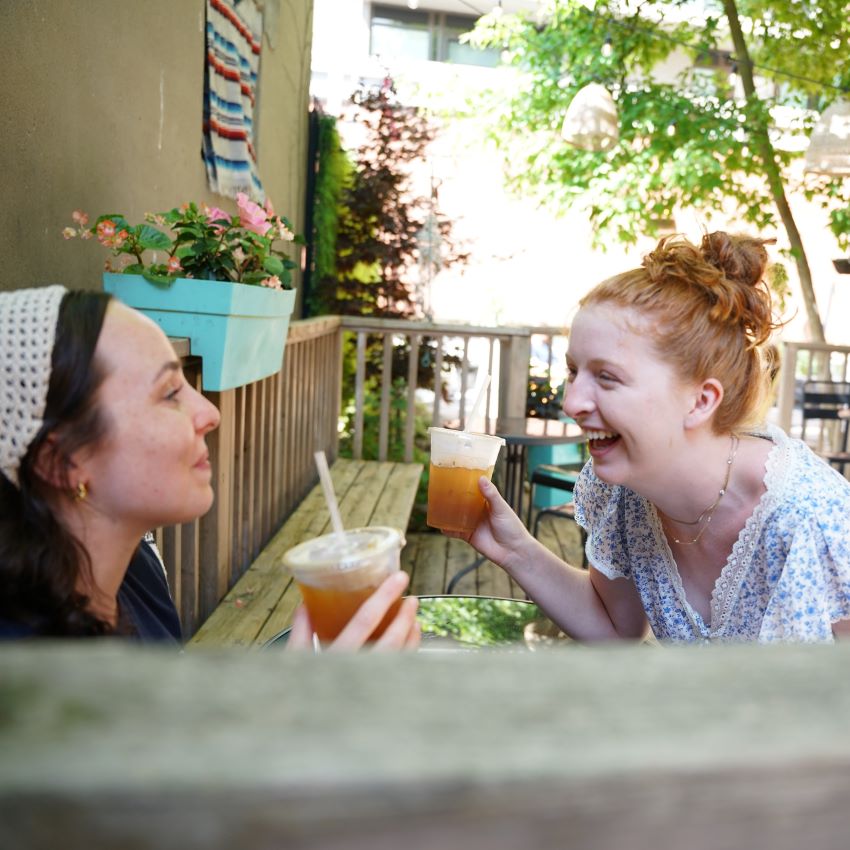 Two women laughing and enjoying drinks on a sunny patio at BSE Skate Shop.