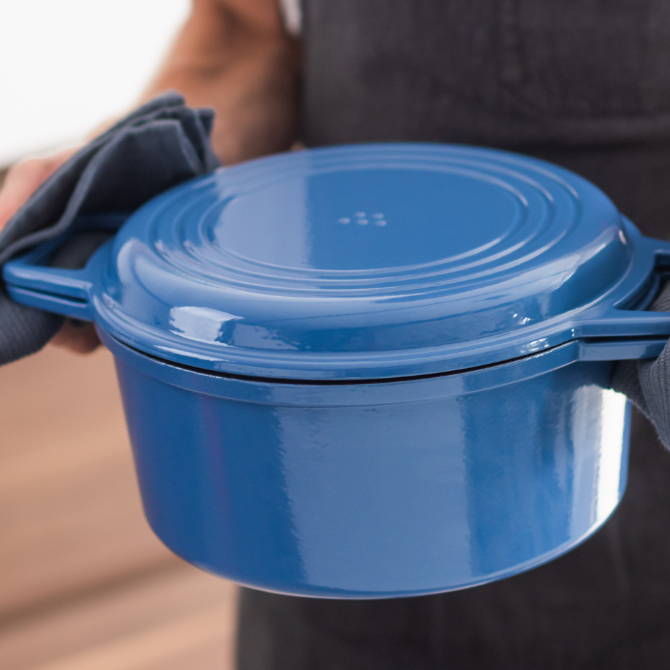A chef carries a Blue Misen Dutch Oven covered with a Grill Lid easily, holding its wide handles with blue kitchen towels.