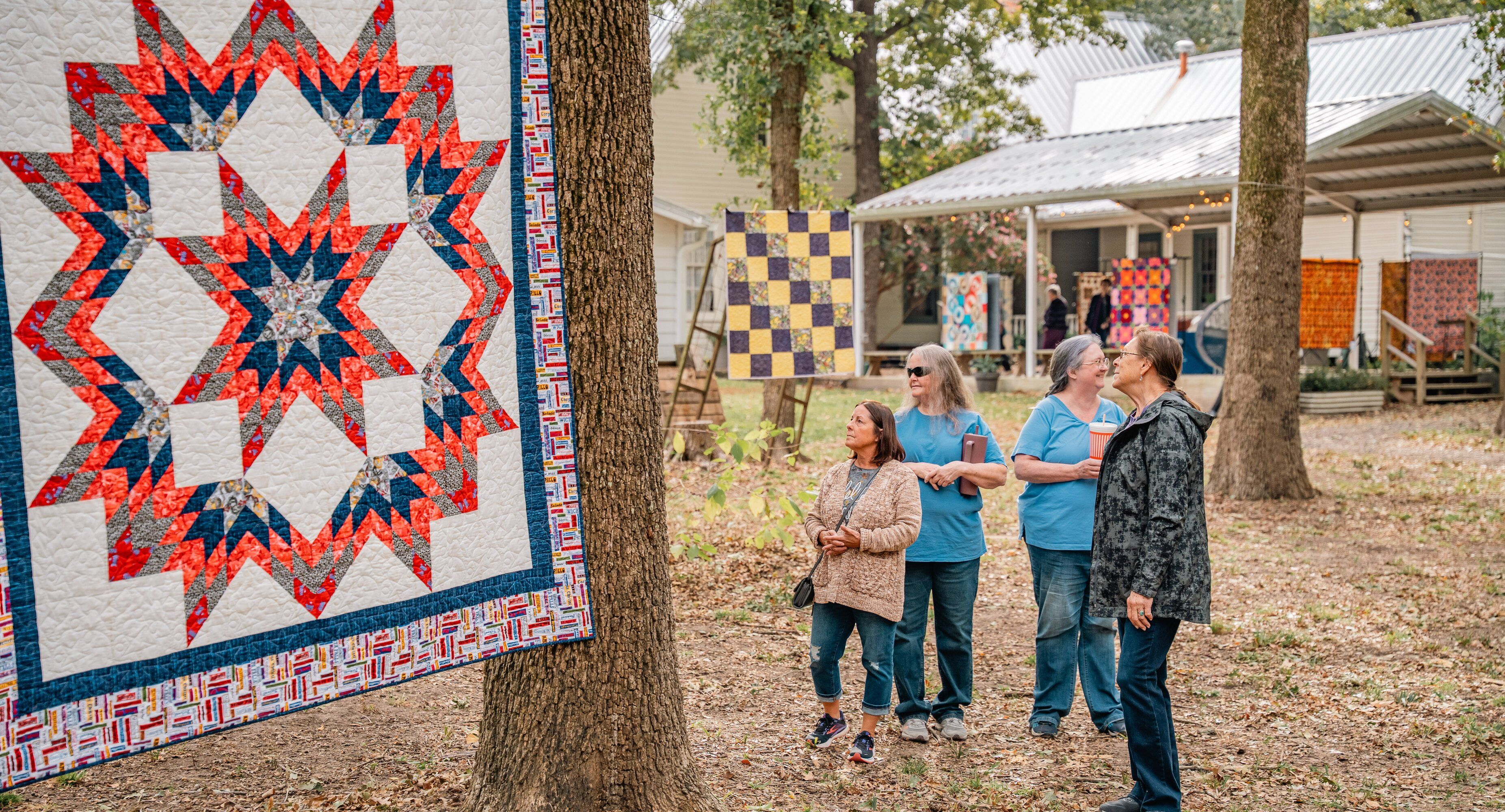 People admiring quilts displayed outdoors near a rustic building.