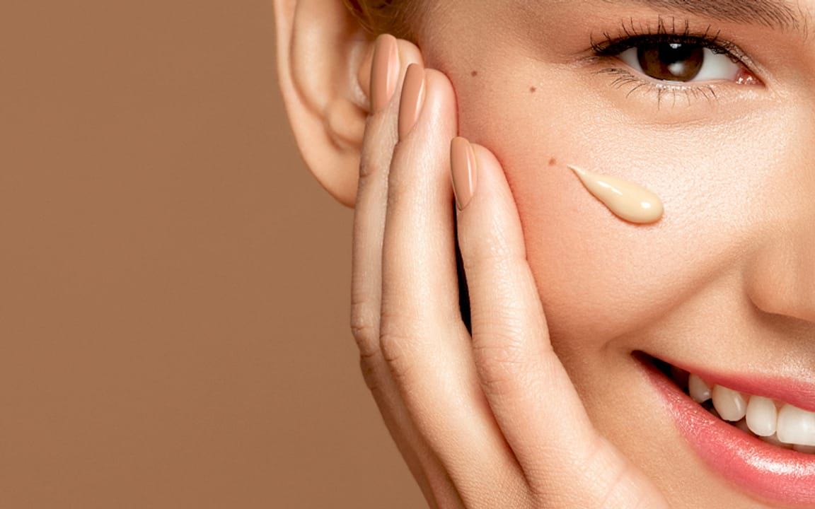 Smiling woman applying cream to her cheek against a neutral background.