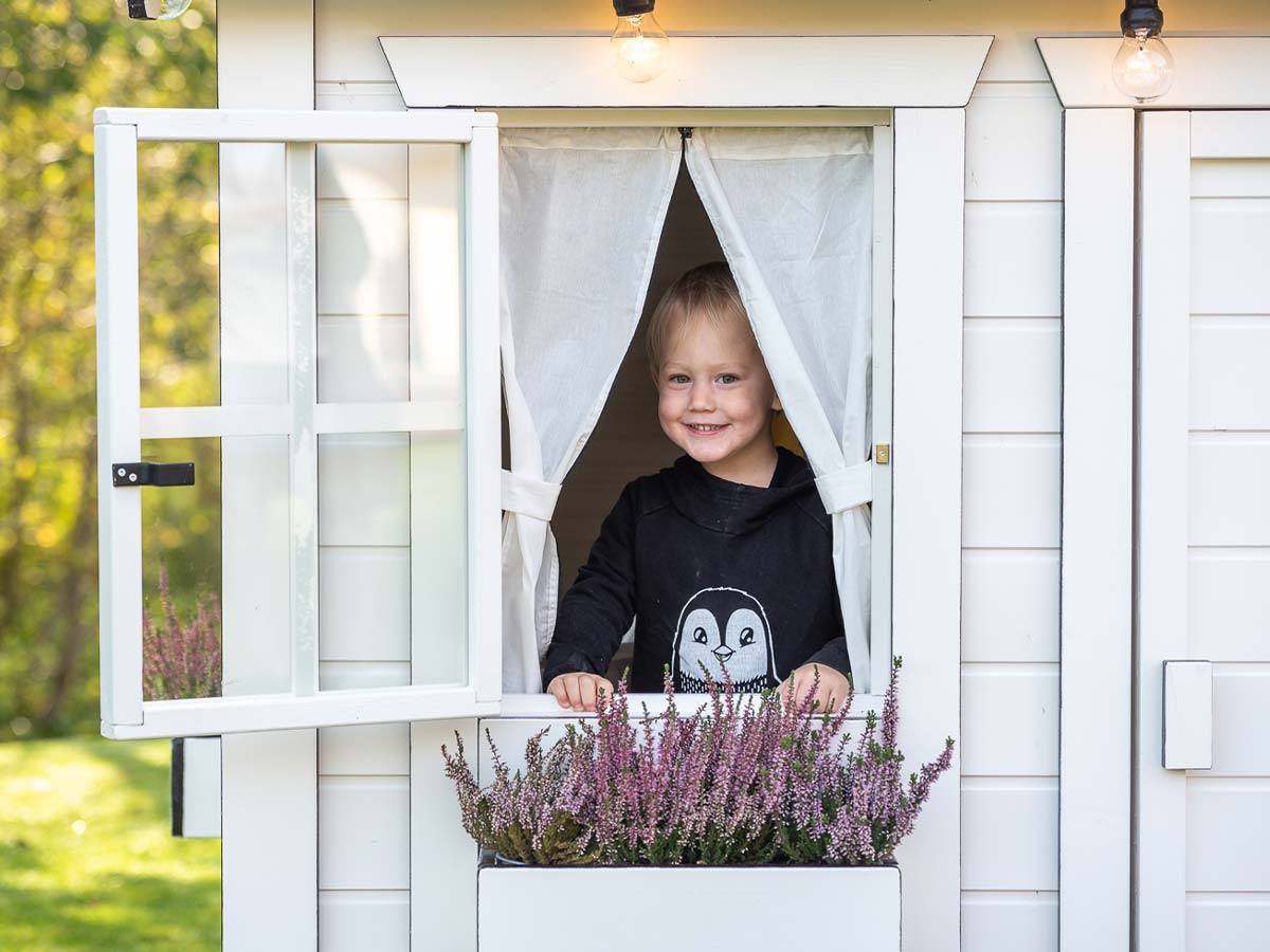 Wooden playhouse with white color with a boy looking out of an open playhouse window by WholeWoodPlayhouses