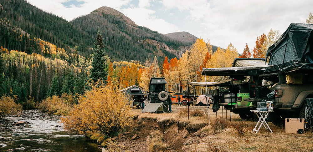 Camping setup beside a river with autumn foliage and mountains in the background.