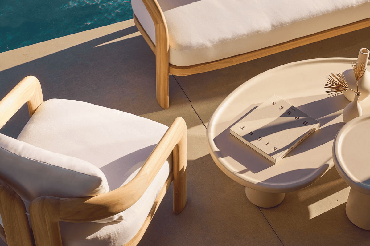 Sunlit poolside seating with a wood-framed lounge chair and white cushion beside two round pedestal side tables holding a book and small vases.