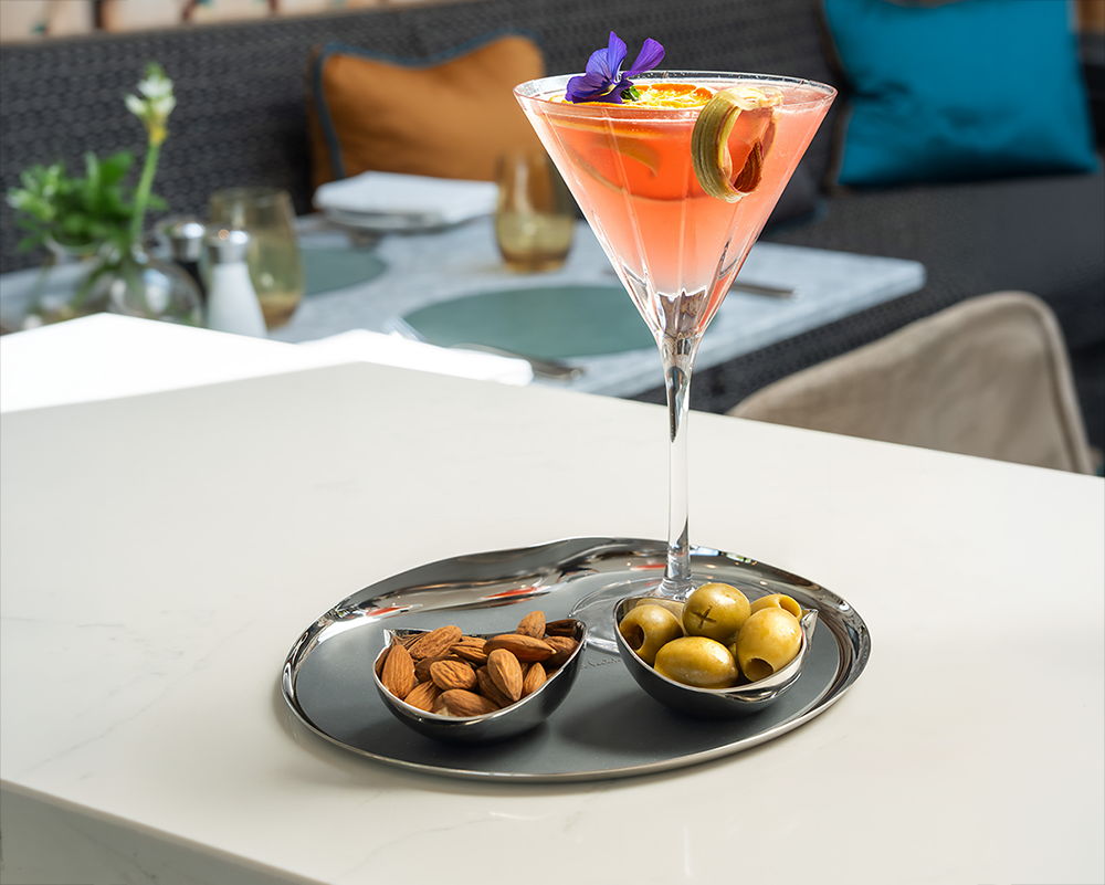 A cocktail with garnishes beside Drift Bowls with almonds and olives on a Drift Serving Tray.
