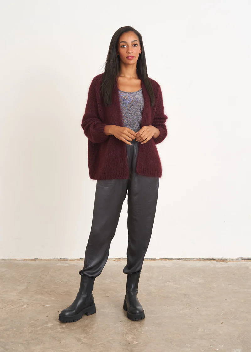 A model wearing a aubergine knitted cardigan over a grey top, dark grey satin trousers and black leather chelsea boots