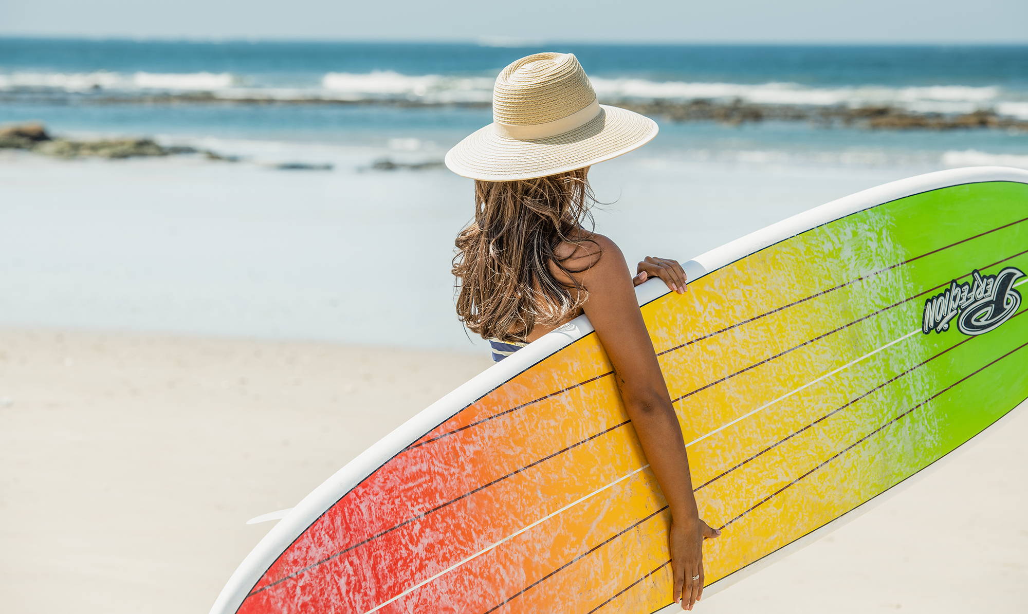 Woman on the beach with a surfboard sporting a Wallaroo Bali, featuring a wide brim and UPF 50+ rating