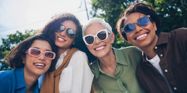 Four women smiling together outdoors, wearing sunglasses in bright sunlight.