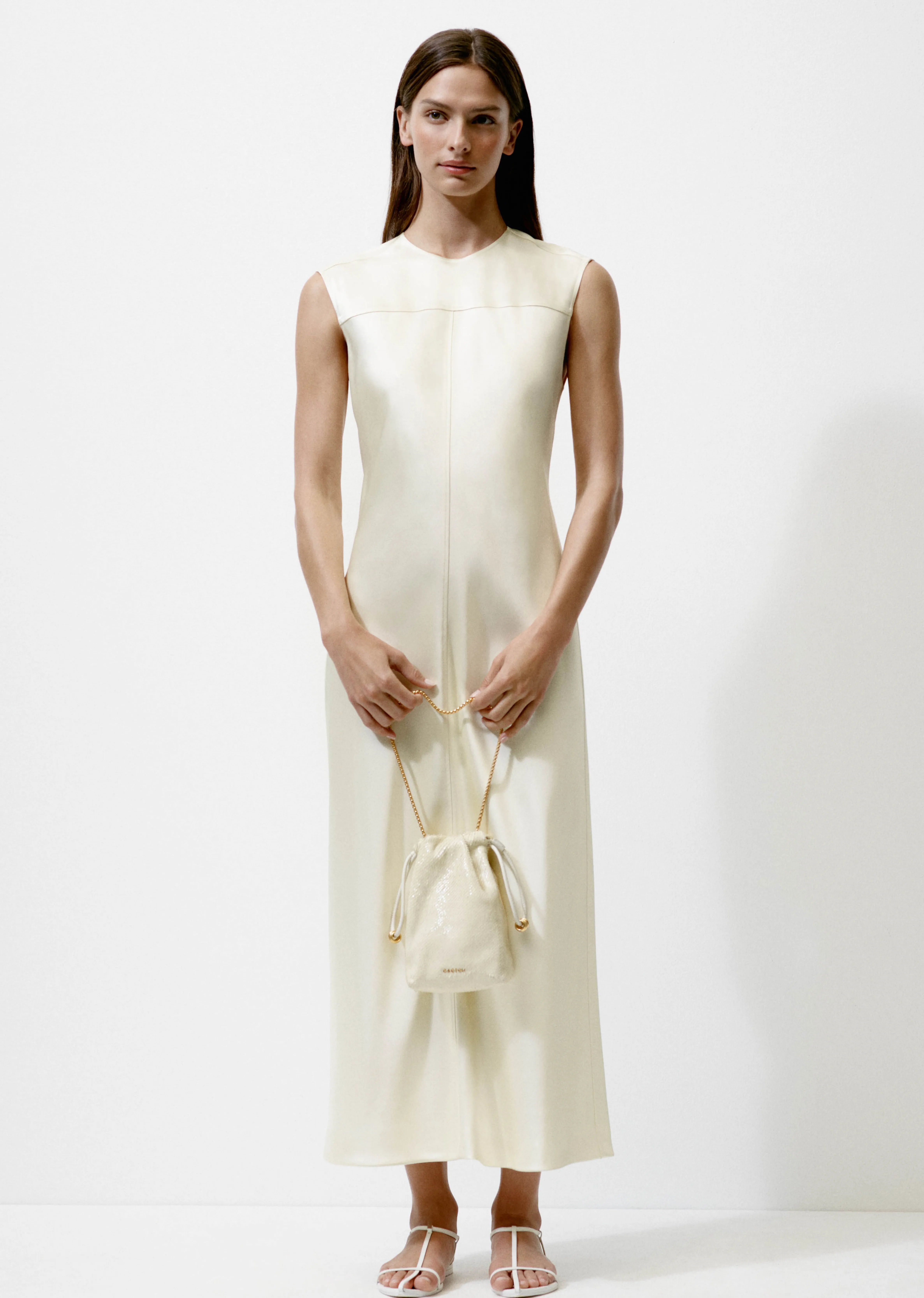 Model wearing a sleek, sleeveless cream dress, holding a small bag.