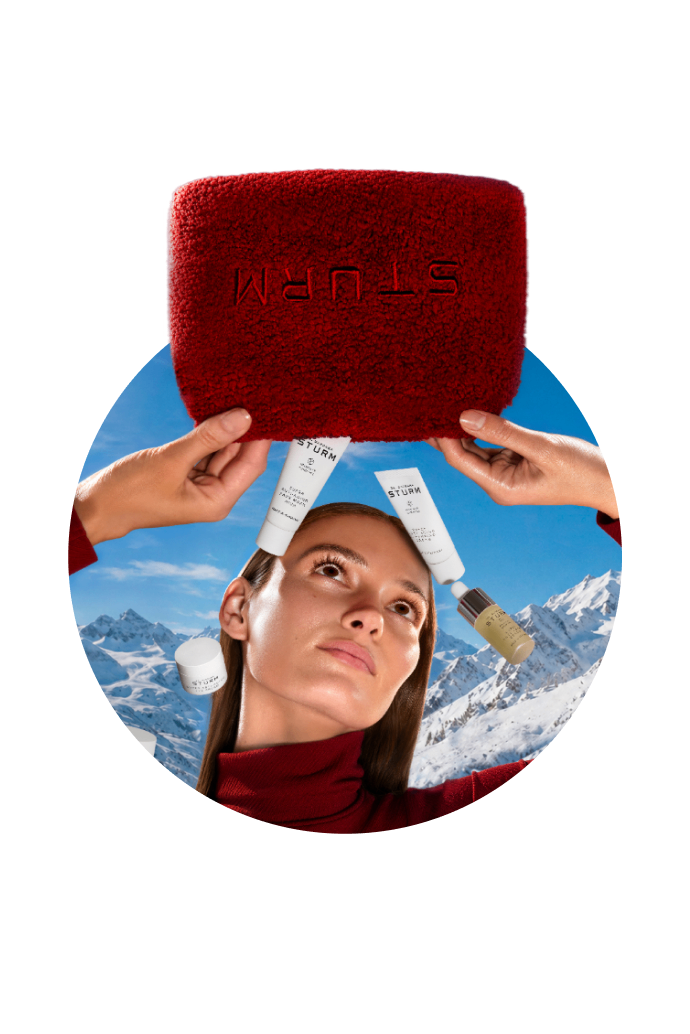 Woman holding skincare products in snowy mountain background, wearing a red sweater.