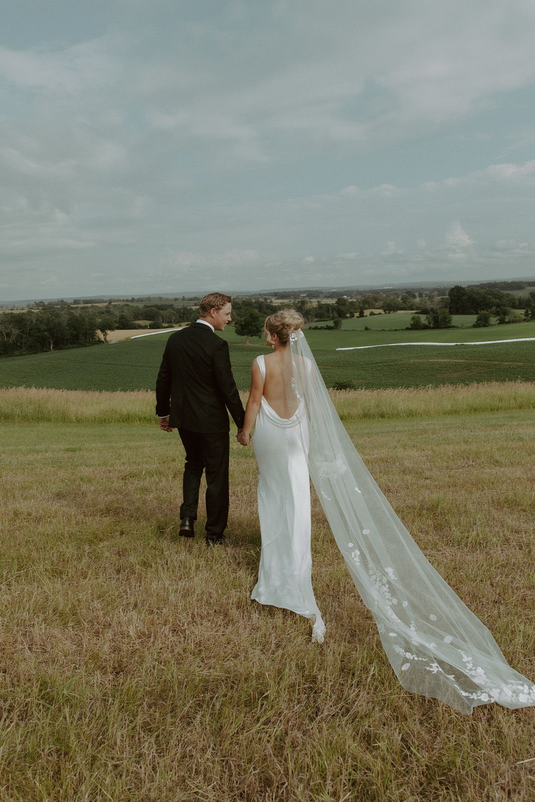 Couple holding hands in a field, bride in white gown and veil.