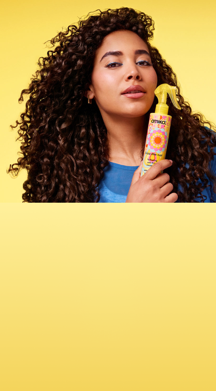 Person with curly hair holding a hair product against a yellow background.