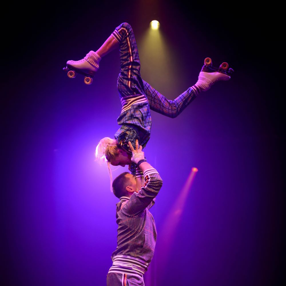 A performer lifts another on roller skates under colorful stage lights.