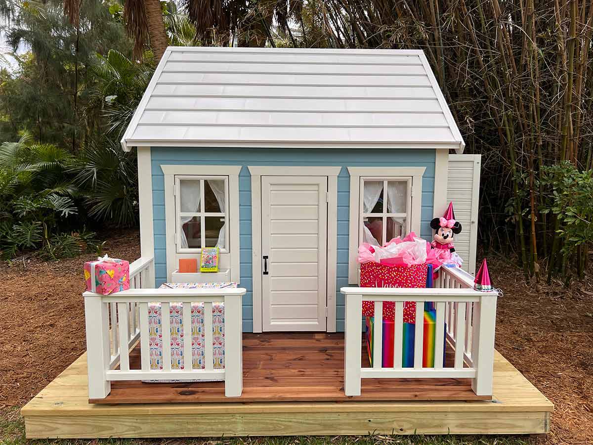 Blue and White Kids Outdoor Playhouse, White Ŕoof, Wooden Terrace with birthday gifts, White Railing, in The BackYard by WholeWoodPlayhouses 