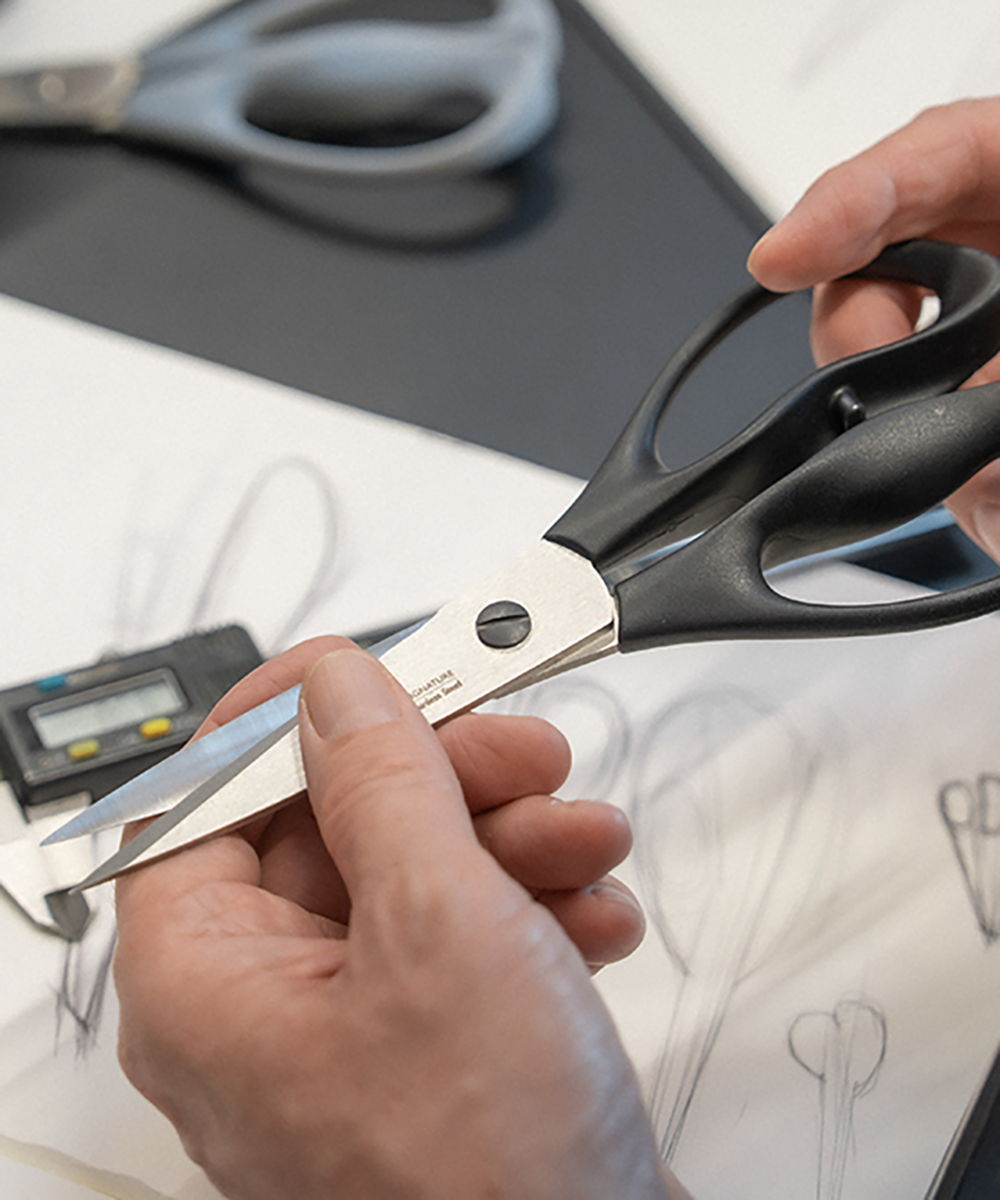 Person holding Signature Scissors over design sketches on a table.