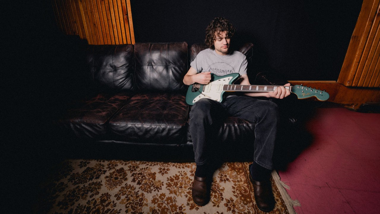 Person sitting on a couch playing a guitar in a studio setting.